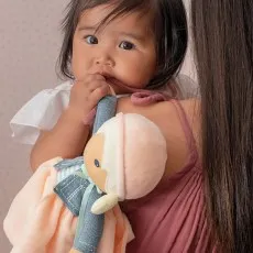 A dark-haired baby, held by an adult wearing a maroon tank top, chews on the arm of a soft Kaloo doll with a pink hat and peach dress..