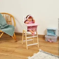 A photo of a doll in a toy high chair, positioned next to a wicker chair in a child's room.