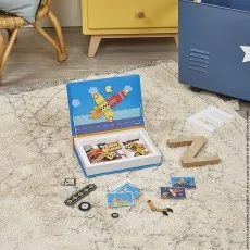 A toy magnetic book kit is open on a fluffy rug, showing a rocket scene inside and various loose magnetic pieces, cards, and wooden letters E and Z scattered around it