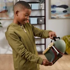 A smiling young boy in a green hoodie holds a Star Wars Boba Fett electronic mask.
