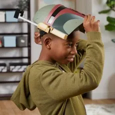 A young boy with a big smile is putting a Boba Fett electronic helmet on his head.