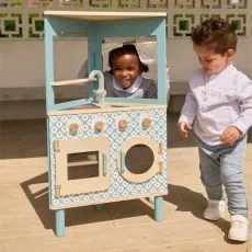 A young girl peeking from behind a light blue and white wooden toy kitchen, with a young boy standing nearby.