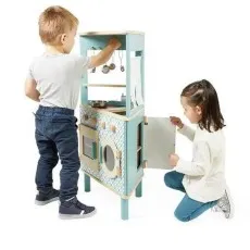 Two children play with a wooden toy kitchen. One child stands while the other kneels, interacting with the set.