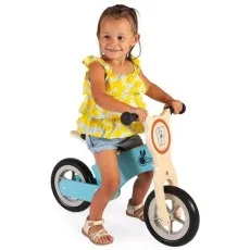 A smiling young girl in a yellow shirt and shorts rides a light blue and wooden balance bike.