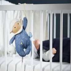 A baby musical plush toy hanging from a white crib's rail, with a sleeping baby in the background