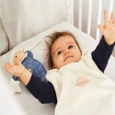 A joyful baby lies in a crib with their arms in the air, next to a plush toy lamb
