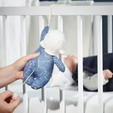 A pair of hands pulls the string of a plush musical lamb toy, hanging from a crib