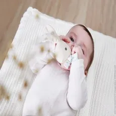 A baby lying on a white knitted blanket, holding a Kaloo rabbit rattle