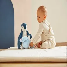 A baby in light-colored overalls sits on a white mat, playing with two Kaloo plush penguins