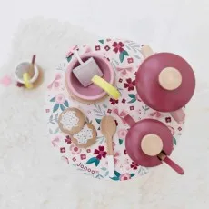 An overhead view of a wooden tea set with a pink teapot, teacups, and two cookies on a floral-patterned mat.