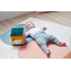 A baby on a patterned rug playing with a multi-textured soft cube toy with a cat illustration.