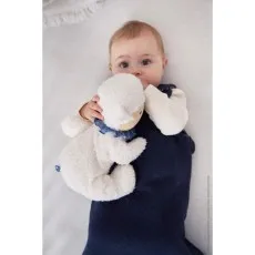 A baby in a dark blue onesie lies on a white blanket, holding and chewing on a soft plush sheep toy.