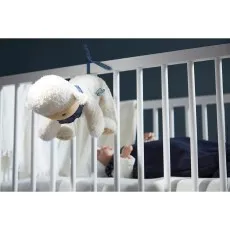 A baby looks up at a hanging plush sheep toy attached to the bars of a white crib.