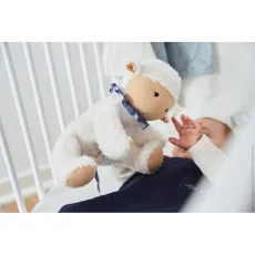 A soft, white stuffed sheep with a blue bow is presented to a baby lying in a crib.