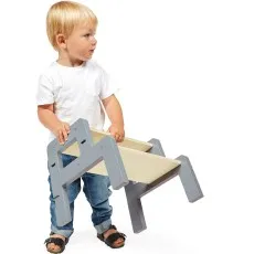 A toddler boy in a white t-shirt and jeans lifts and carries a small wooden step stool.