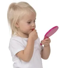 A toddler mimics putting on makeup, holding a toy lipstick and a matching pink mirror.