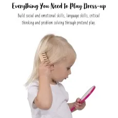 A young blond-haired child holds a wooden toy comb to her hair while looking into a pink toy mirror.