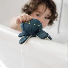 A child's hand holds a dark blue octopus bath toy over the edge of a white bathtub.