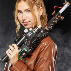 A young woman in a brown leather jacket holds a Ghostbusters Proton Pack wand over her shoulder