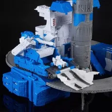 A close-up of a Transformers playset showing a blue tank with a small white vehicle on its ramp