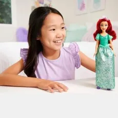 A young girl with a broad smile looking at a red-haired Ariel doll on a table.