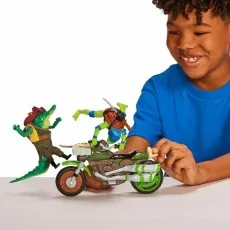 A smiling boy in a blue shirt is playing with his Teenage Mutant Ninja Turtles action figures and a motorcycle.
