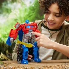 A smiling child plays with the Optimus Prime action figure, pressing a button on its chest, with a blurred natural background