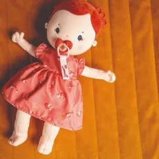 A doll with red hair and a pink-red dress is lying on a soft, orange blanket