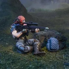A G.I. Joe Classified Series Cobra Zandar action figure sitting on the ground, aiming a rifle with a scope. His backpack sits next to him