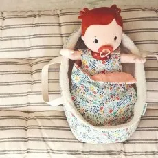 A red-haired cloth doll with a pacifier lying in a floral-print, sherpa-lined toy carrycot on a striped cushion