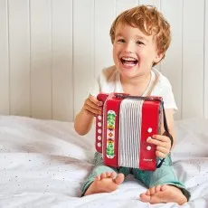 A joyful young child sitting on a bed, laughing while holding a red Janod toy accordion decorated with flowers