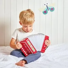 A young boy sitting on a bed, focused intently on playing a red Janod toy accordion with flower art
