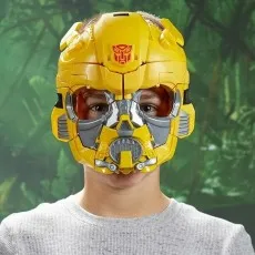 Close-up of a child wearing a detailed, yellow Bumblebee robot mask with gray facial details against a green jungle background..