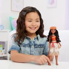 A smiling girl in a denim shirt plays with her Disney Princess Moana doll indoors.
