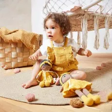 Toddler in a yellow romper sitting on a rug, playing with two Kaloo yellow dress rag dolls.